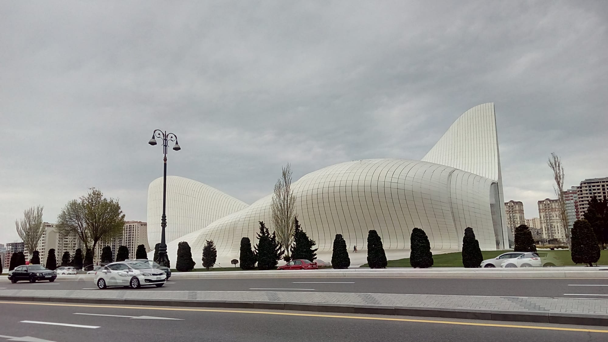 A large white building of flowing curves rising to a swept-back peak, seen across a wide road with cars and a row of clipped dark conifers