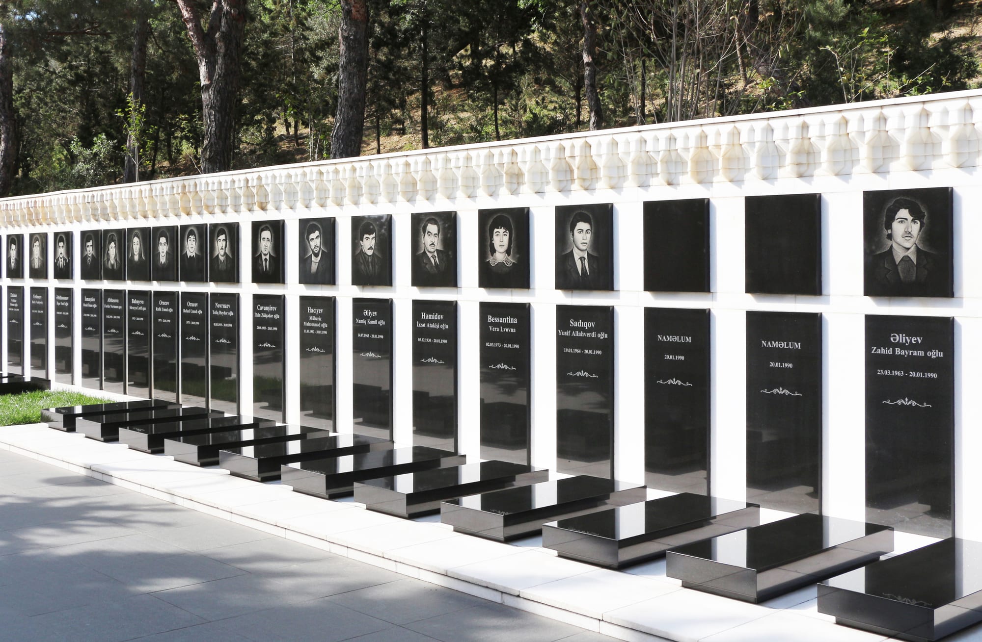Rows of black polished gravestones bearing portraits along a landscaped hilltop terrace overlooking a bay