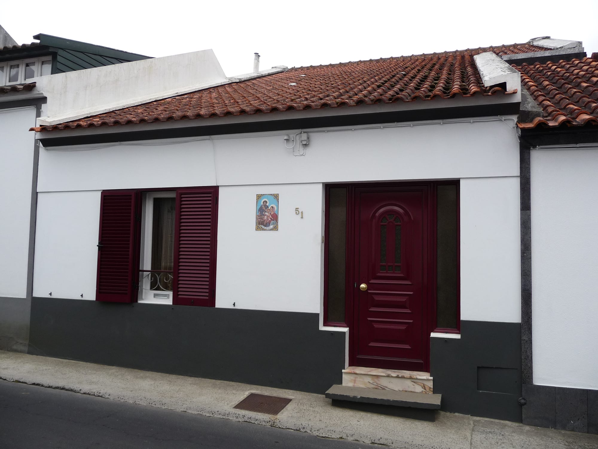 A single-storey white house with a red front door, red-shuttered window and terracotta tile roof, on a village street