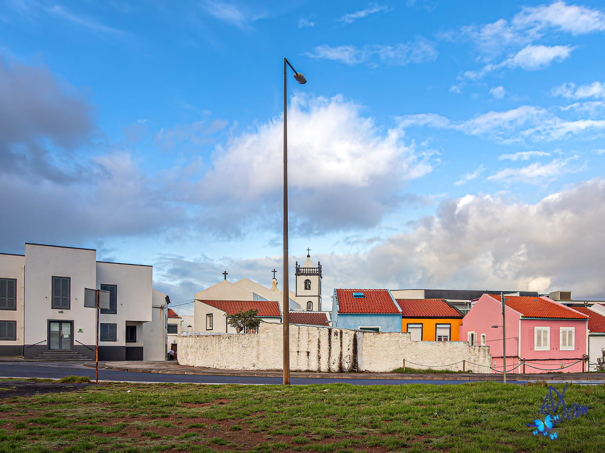 A residential street on São Miguel with a parish church tower and a row of houses painted white, blue, orange and pink, under a partly cloudy sky