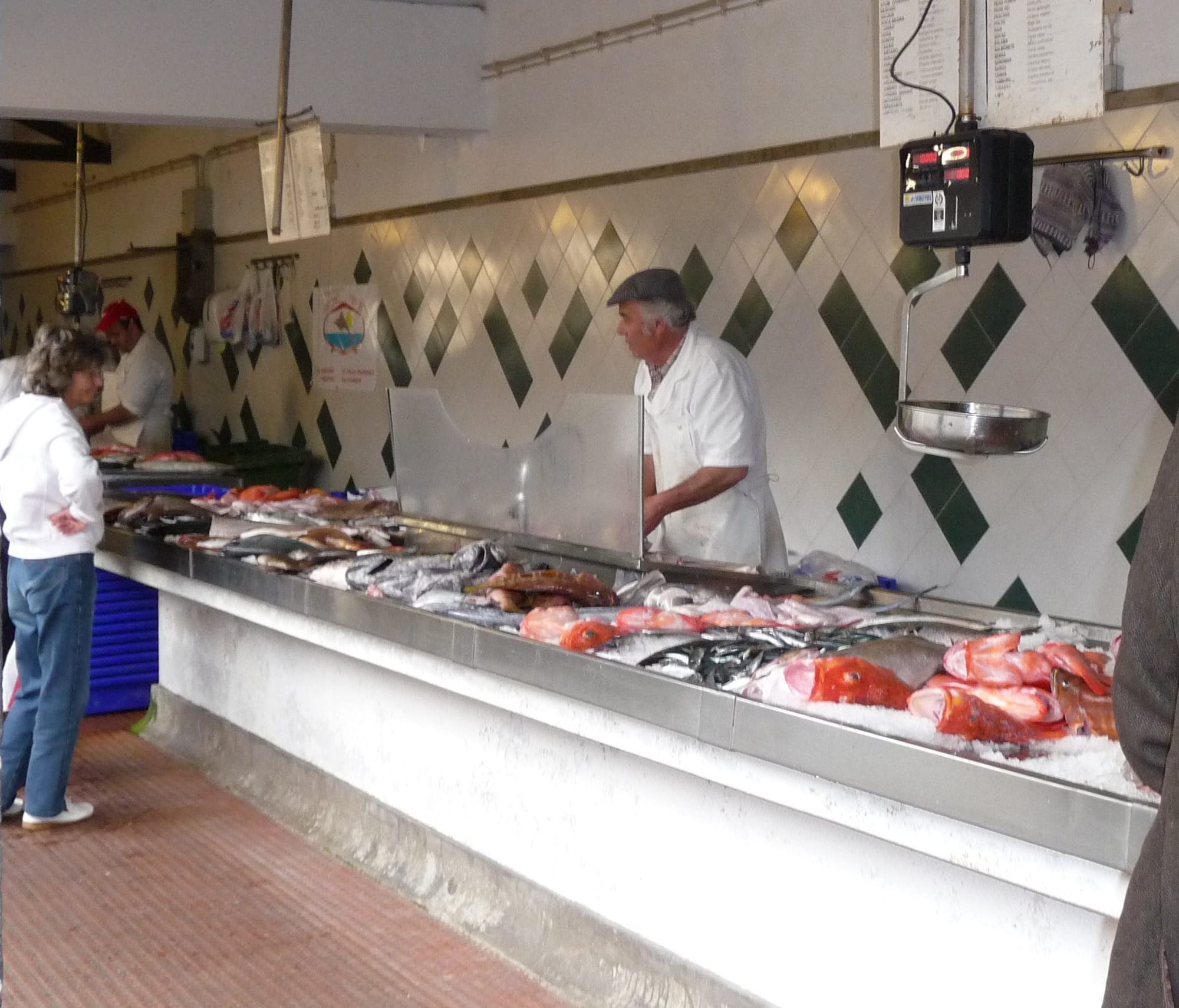 A fish vendor in a white coat serving a customer at a market stall piled with fresh fish on ice, patterned tiles on the wall behind