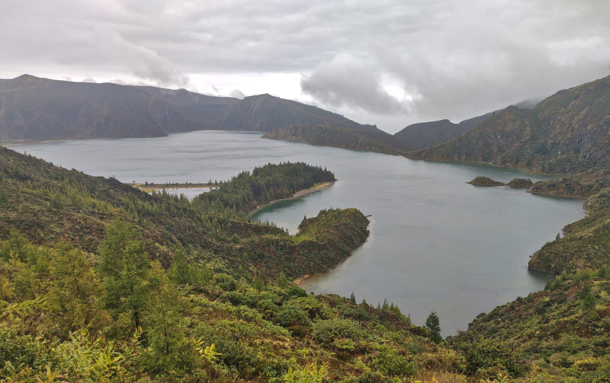 A blue crater lake with a sandy shore, ringed by steep green slopes often in cloud