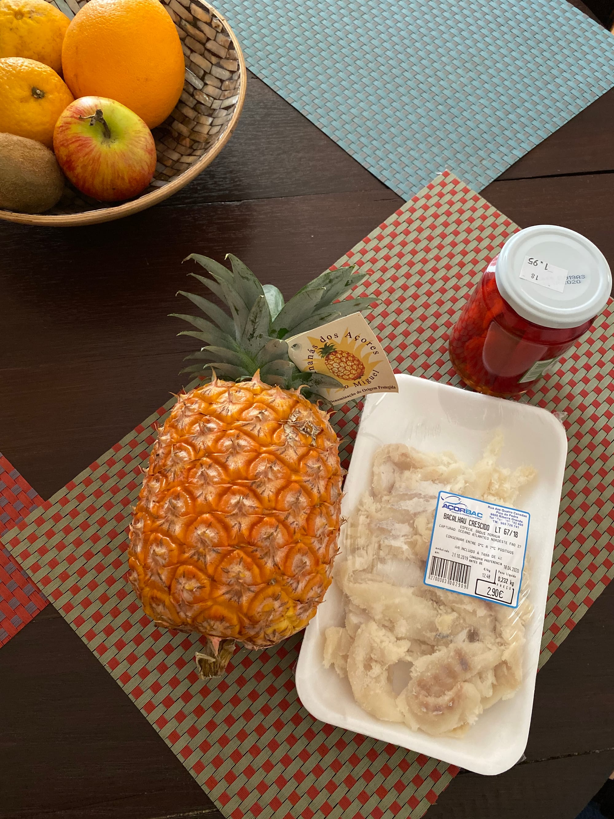 A small pineapple with a producer's tag reading 'Ananá dos Açores' on a table beside fresh fruit and salt cod