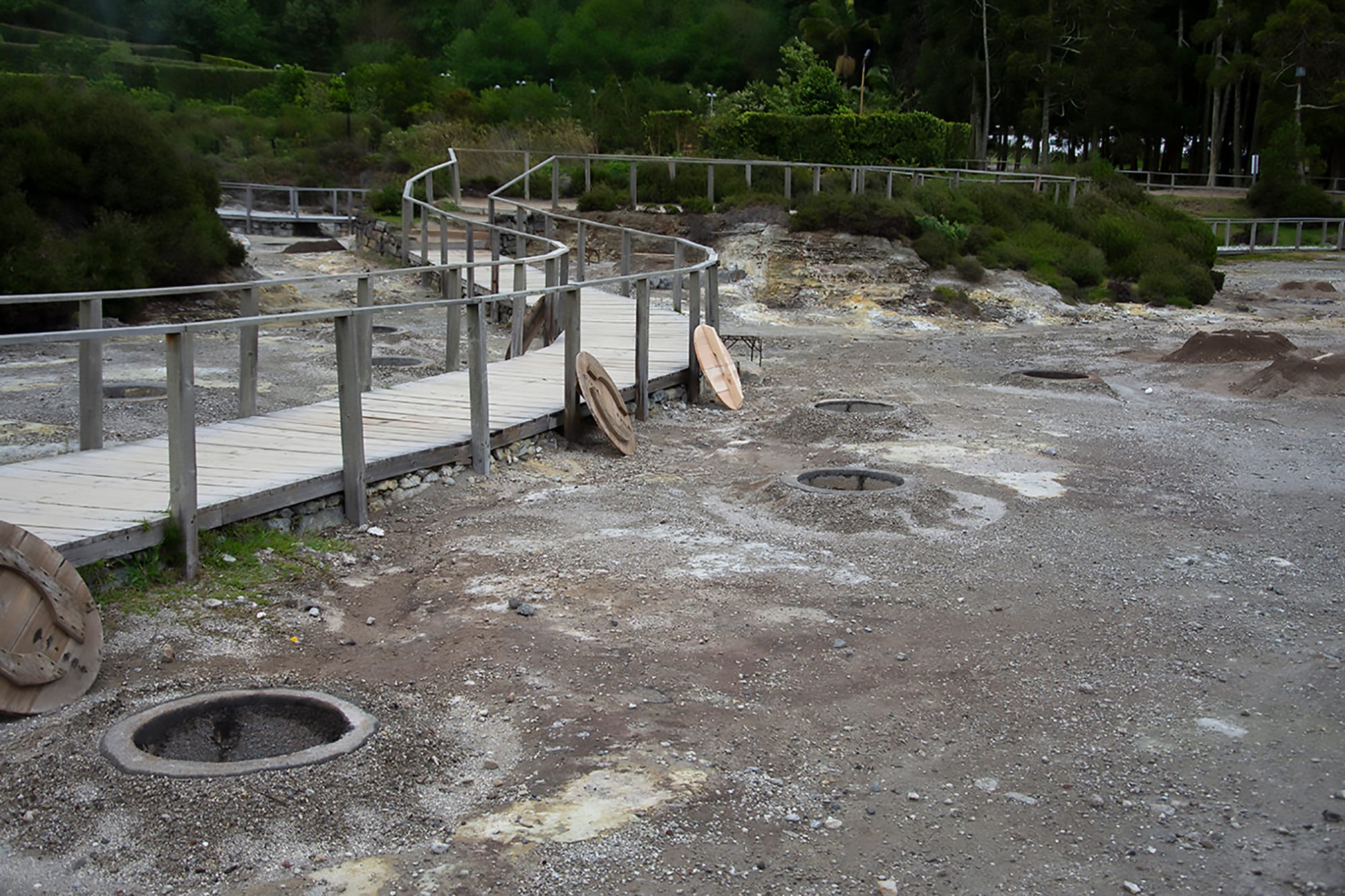 A boardwalk running past steaming volcanic vents and covered cooking pits set into bare, mineral-stained ground