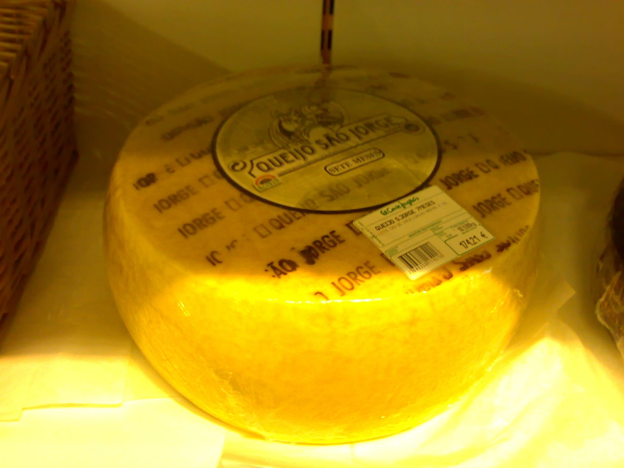A whole wheel of pale yellow cheese with its label visible, on a shop shelf