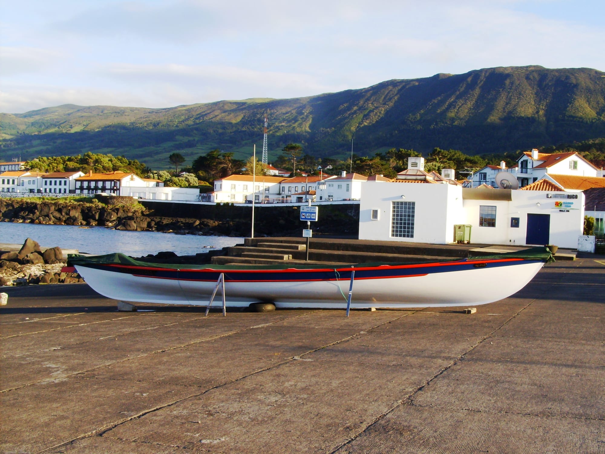 A long, slender white open boat with a red and blue stripe propped on a stone quay, with white harbour houses and green hills behind