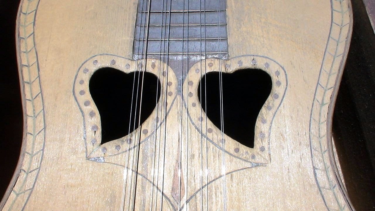 Close-up of the pale wooden face of a guitar with two heart-shaped soundholes side by side, outlined with small dots, and a set of steel strings running over them