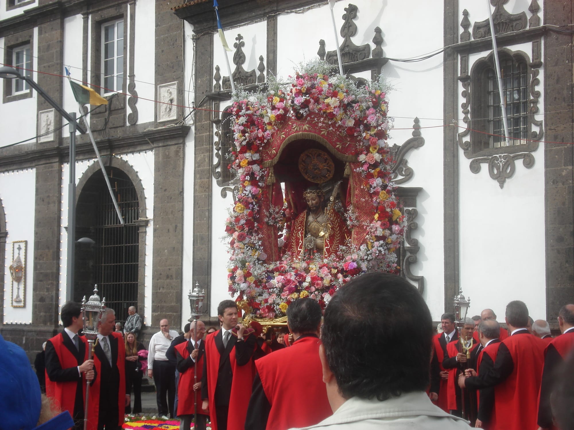 An image of Christ in a red robe framed by a large arch of pink, red and yellow flowers is carried out of a church, surrounded by men in red capes holding lanterns on poles