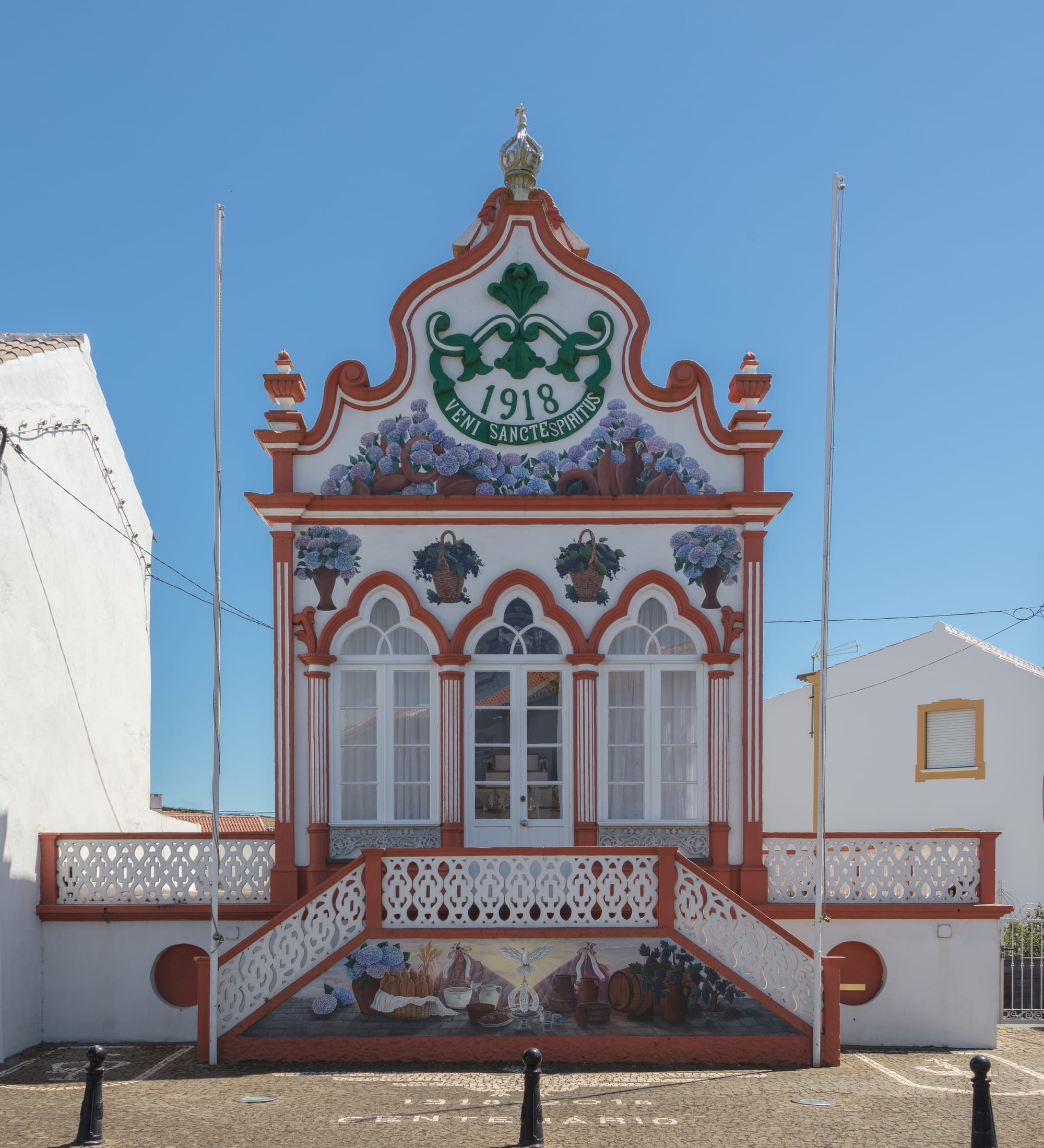 A small white chapel trimmed in terracotta red, crowned with a silver crown, its gable painted with hydrangeas and the words Veni Sancte Spiritus, with a decorated staircase in front