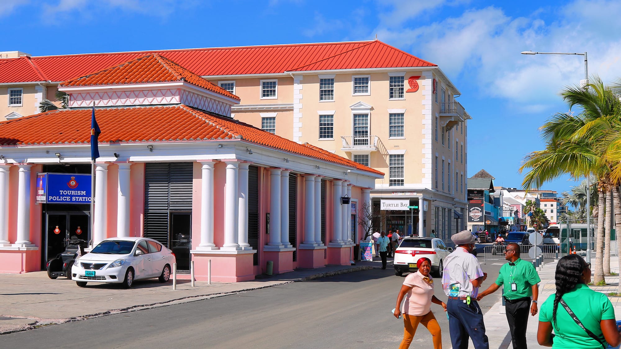 A pink and cream colonial-style building with a red tiled roof on a Nassau street, with several people crossing the road in the foreground