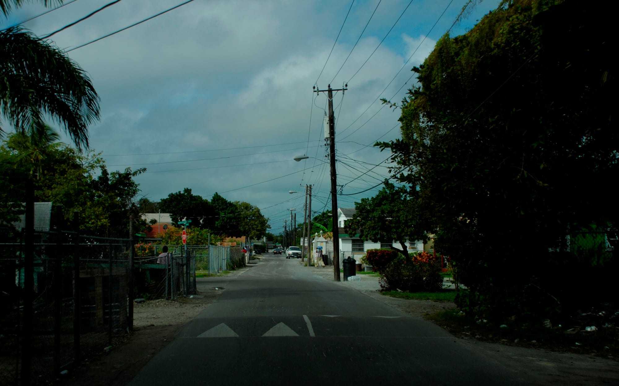 A quiet residential street in Nassau lined with small houses and palm trees, with power lines overhead and one person walking along the fence line