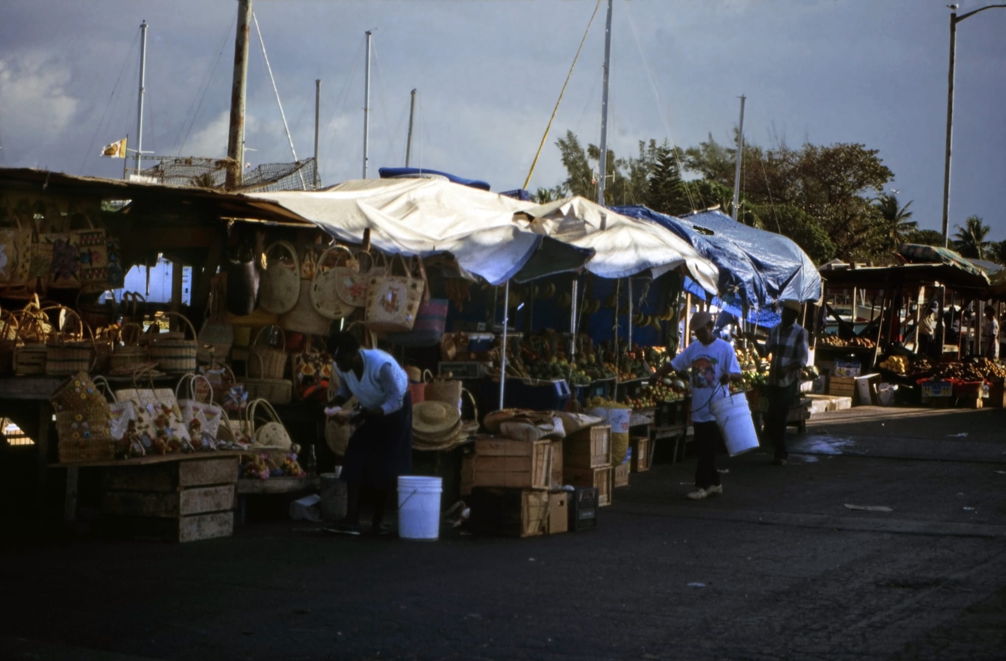 Vendors and a shopper at an open-air market stall selling straw baskets, hats and produce under tarpaulin awnings at a dock