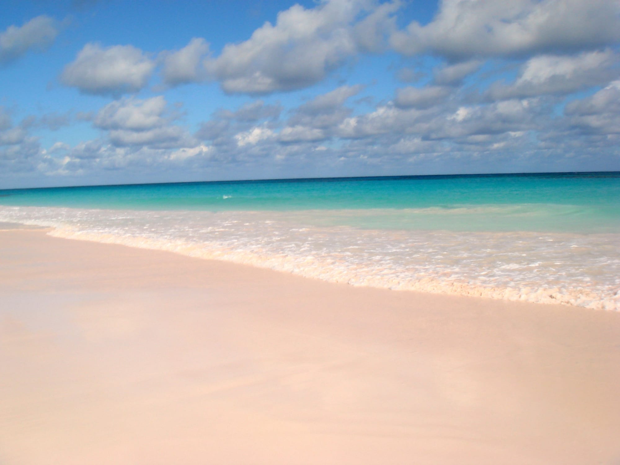 A long beach of pale pink sand meeting clear shallow water under a bright sky