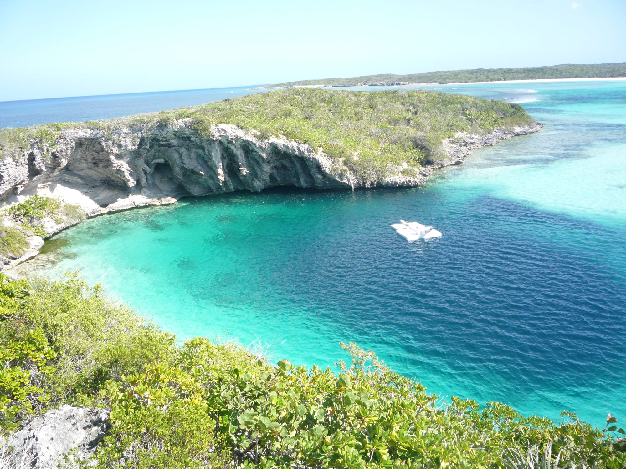 A near-circular pool of dark blue water against a turquoise lagoon and low cliffs