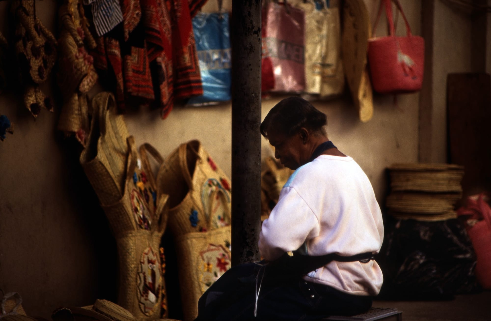 A woman in a white sweatshirt sits working with her head bowed beside a stall hung with woven straw bags, hats and coloured fabric