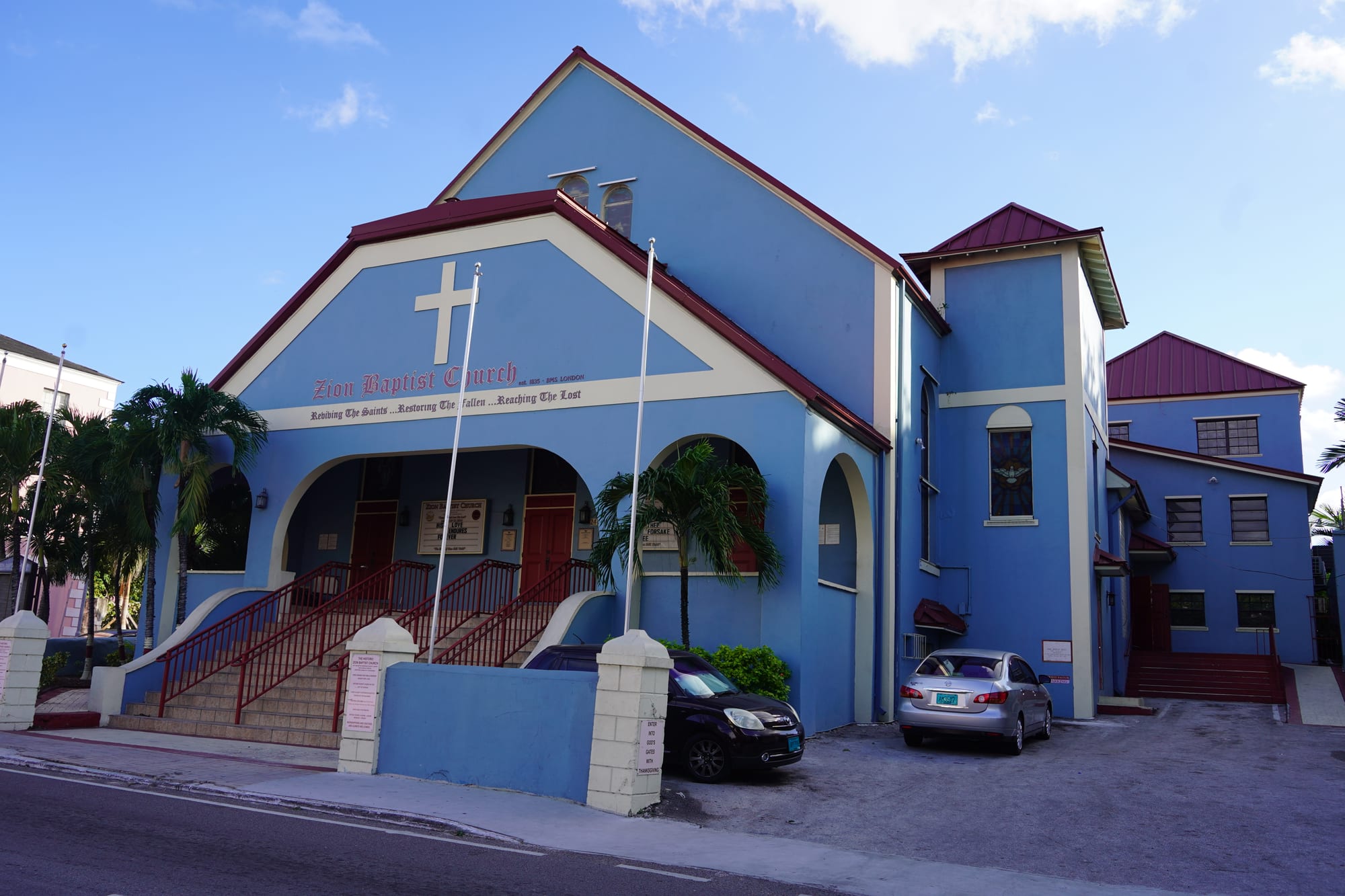 A large pale-blue church with a white cross on its gable, a red roof and twin staircases with red railings leading up to an arched entrance, beside palm trees