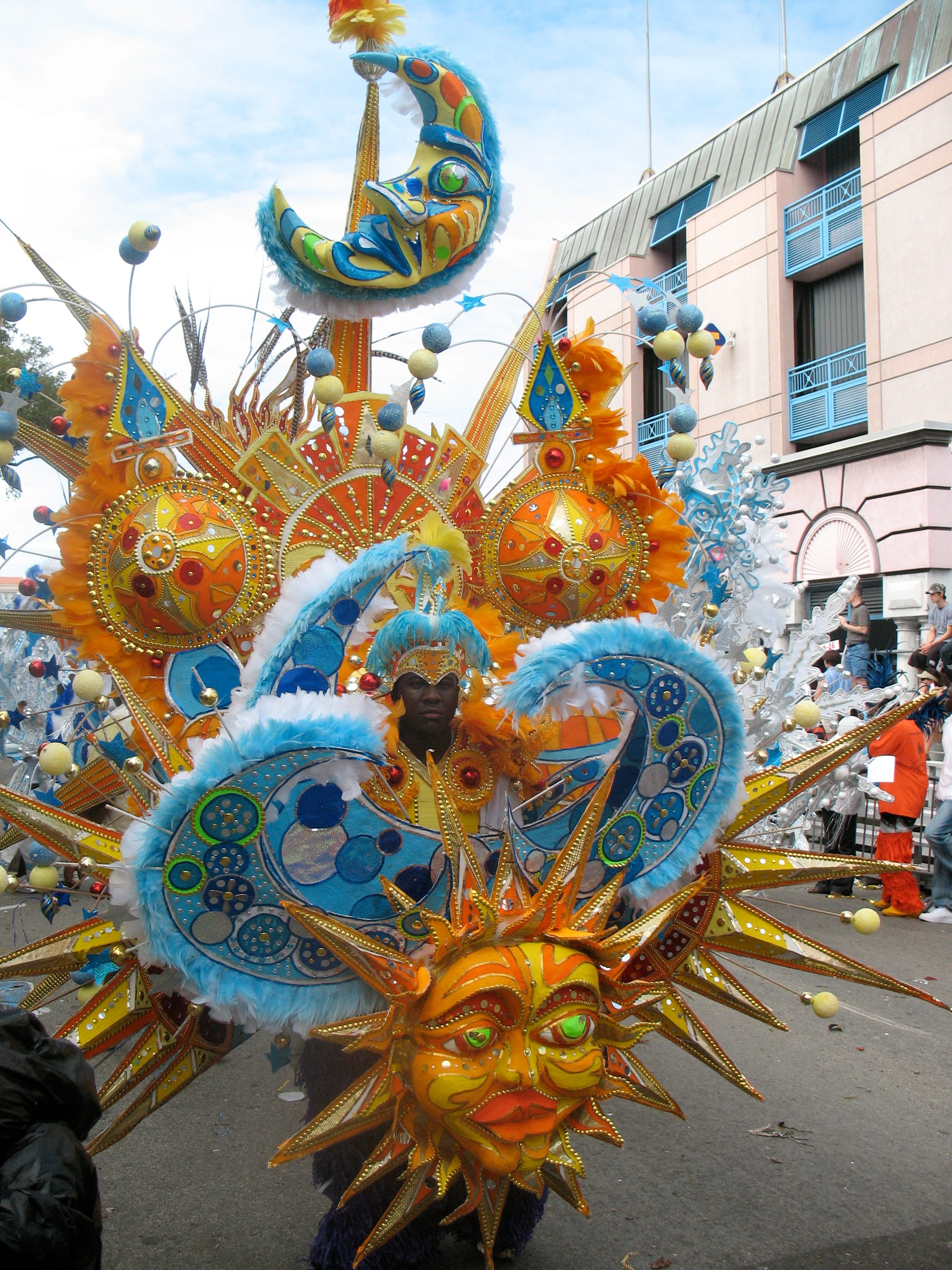A Junkanoo performer in a huge costume of golden suns, a crescent moon and blue feathered swirls, standing in a street with spectators on a balcony behind