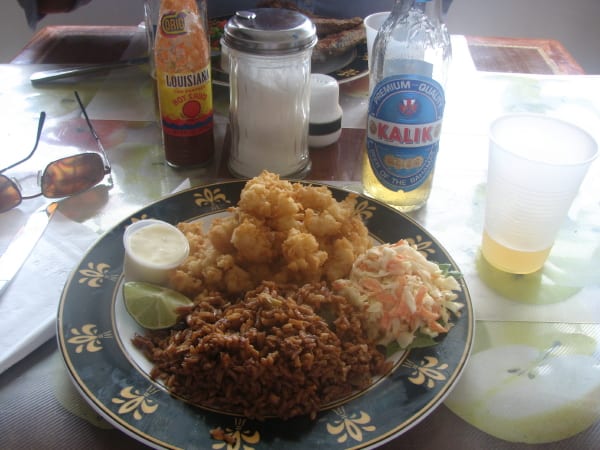 A plate of golden fried conch fritters, brown pigeon-pea rice and coleslaw, with a bottle of Kalik beer behind it