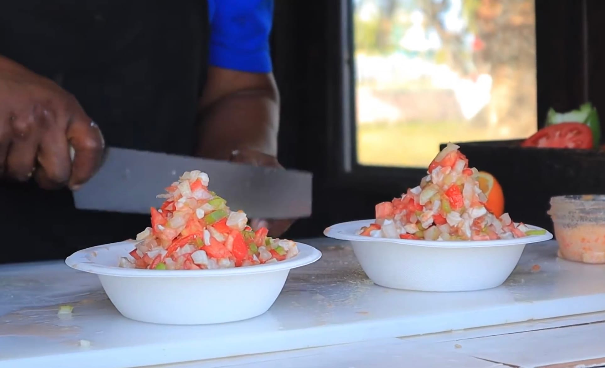 A vendor's hand chopping diced conch salad with a large knife, with two heaped bowls of the salad in front