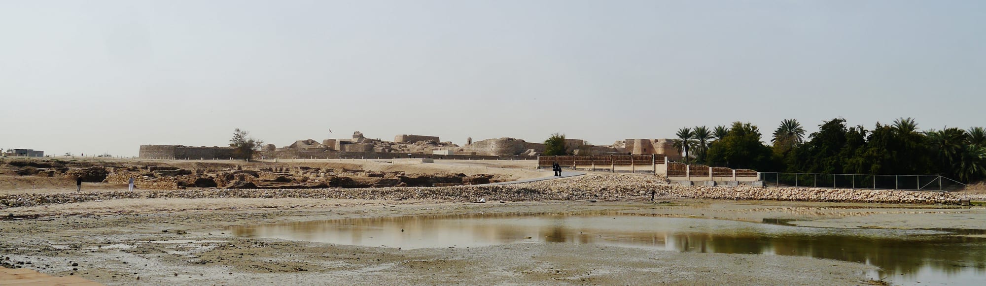 Excavated stone foundations and walls of a layered ancient site beside a modern fort