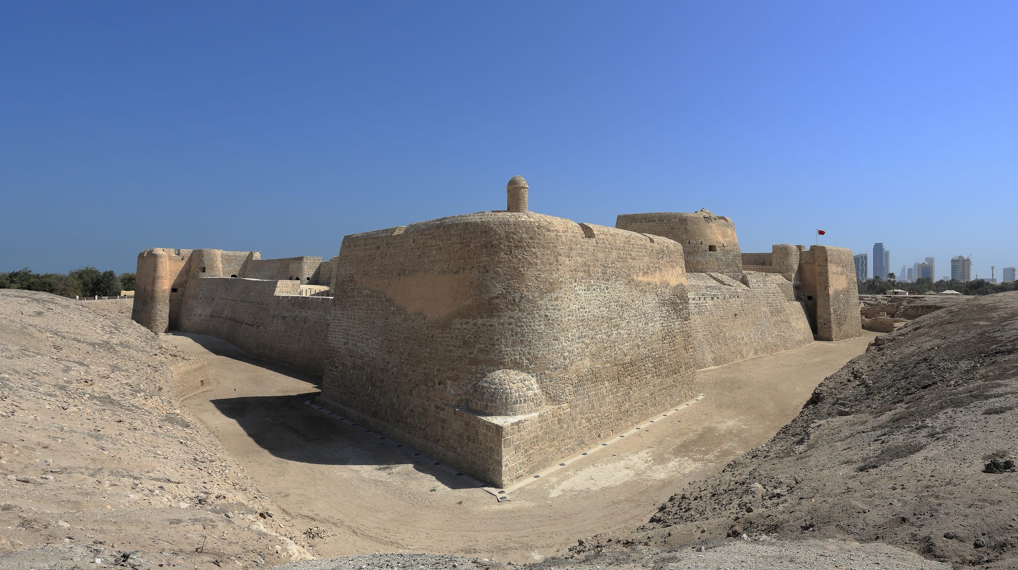 A low sand-coloured fortress with square towers on a rise, palm trees around it