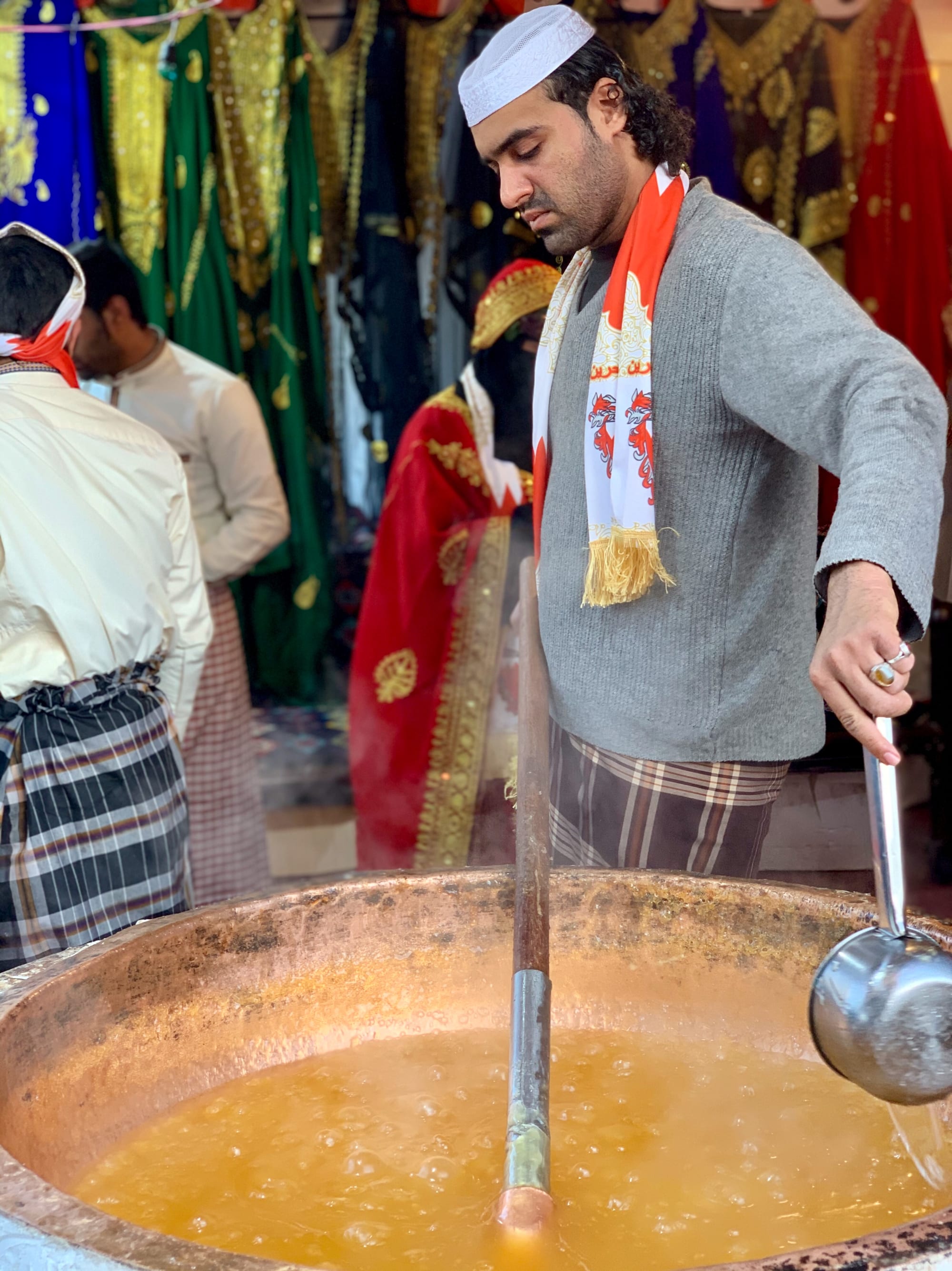 A person stirring a large pan of dark, glossy halwa over an open flame at an outdoor festival