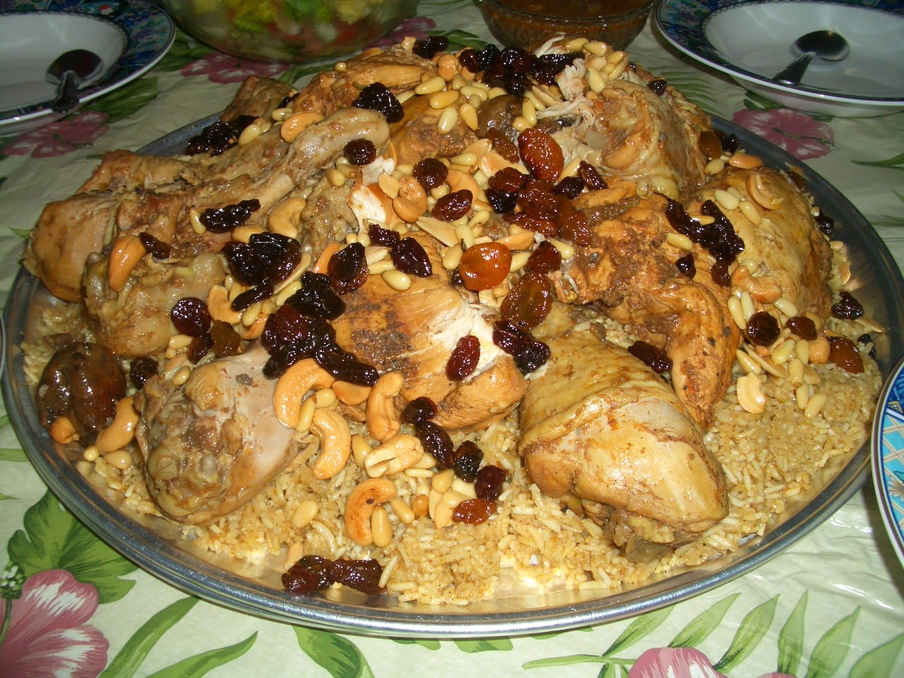 A large round metal platter of spiced rice piled with pieces of chicken and scattered with cashews, pine nuts and dark raisins, on a flowered tablecloth