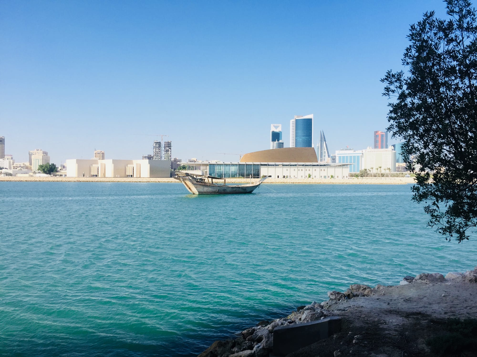 A weathered wooden dhow moored in turquoise water, with low white museum buildings, a gold-roofed theatre and glass towers on the far shore