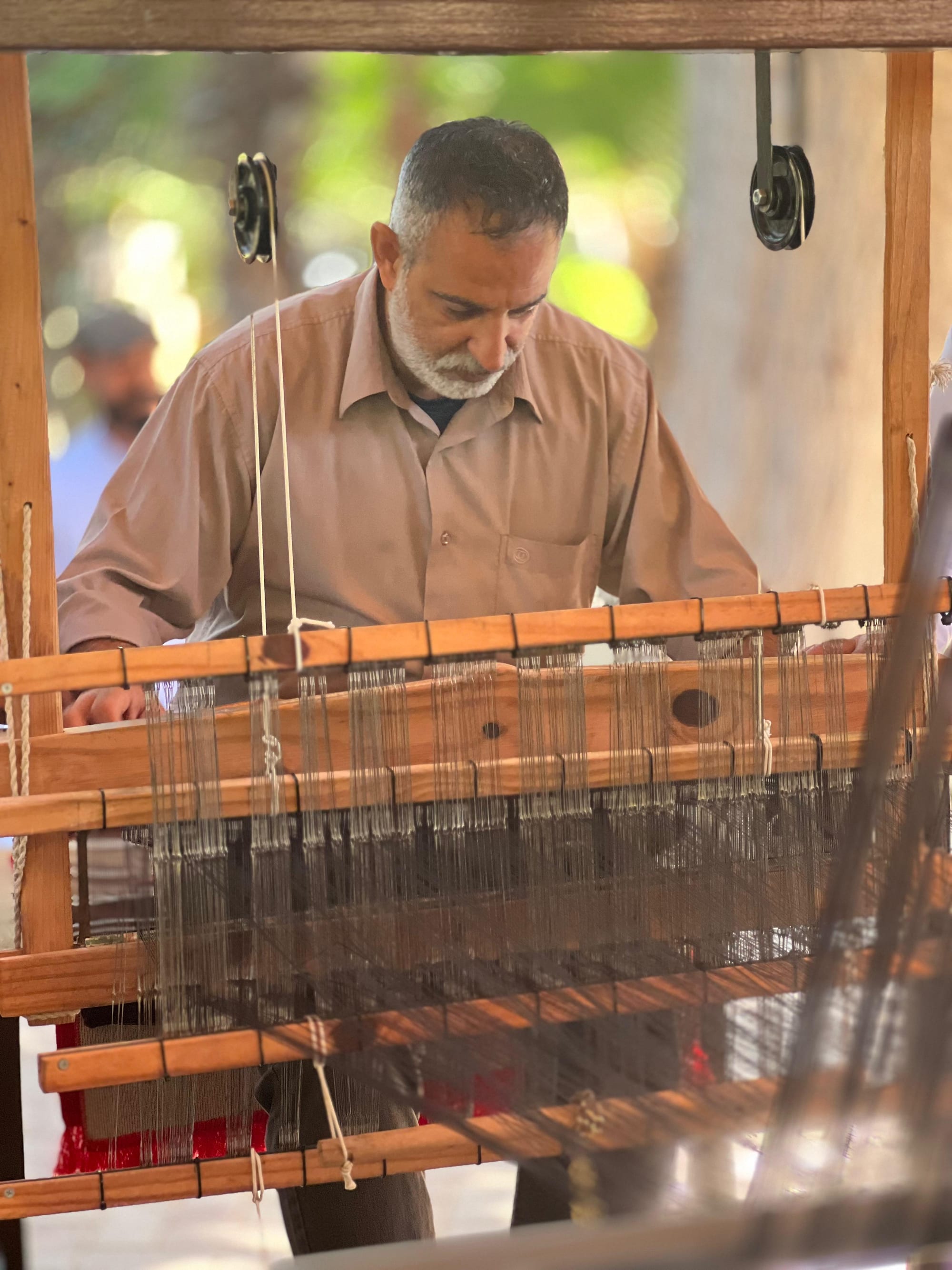 A grey-bearded man in a beige shirt works at a large wooden hand loom strung with fine threads, seen through the loom's frame
