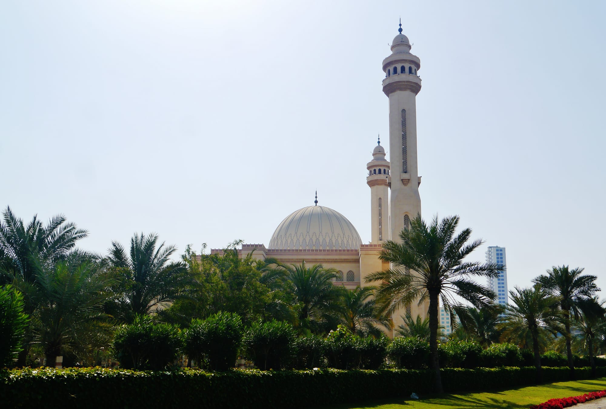 A large cream-coloured mosque with a ribbed dome and two slender minarets rising above palm trees and a clipped hedge, under a pale sky