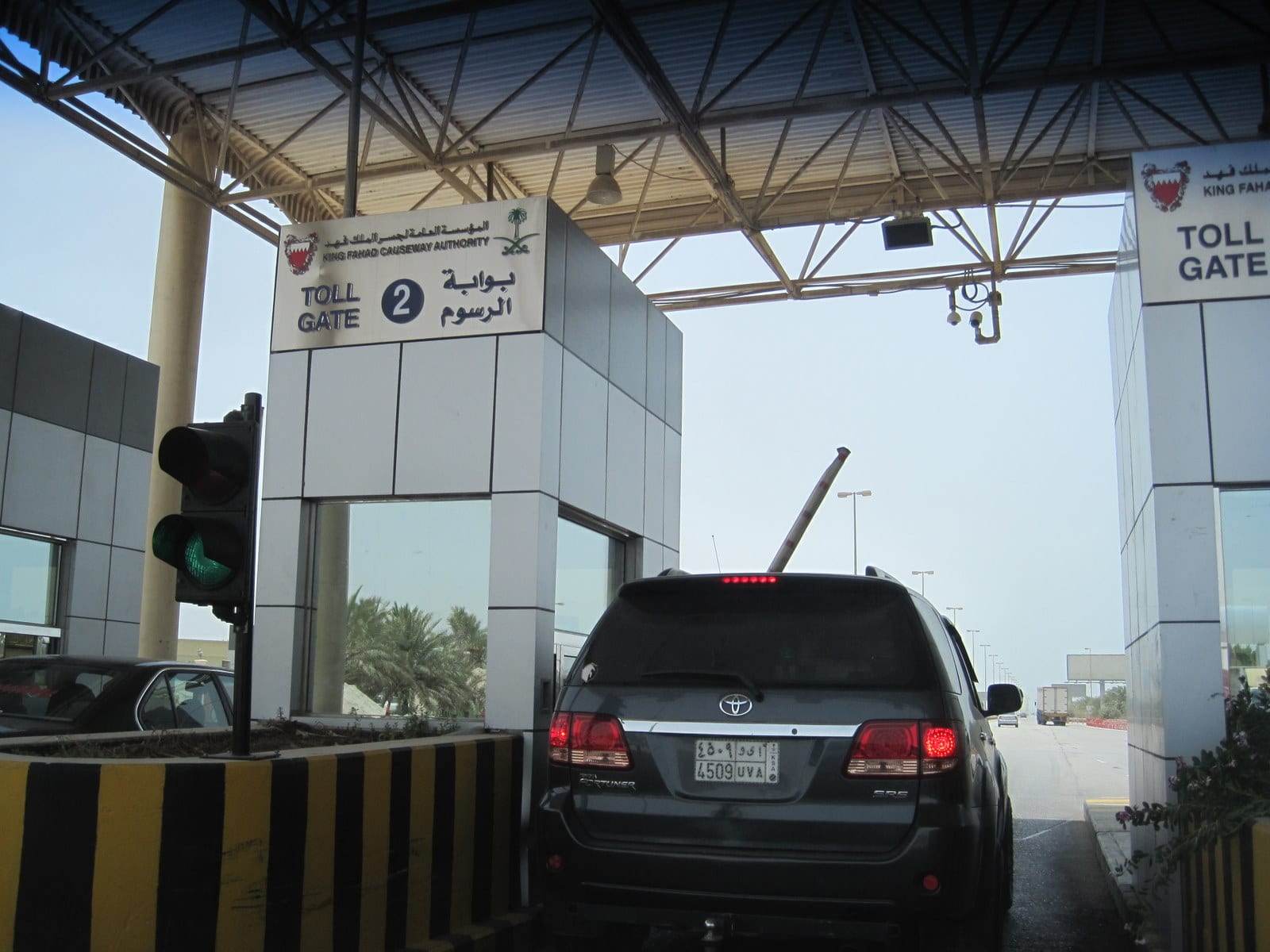 A car queuing at a King Fahd Causeway toll gate booth, with signage in English and Arabic and another lane of traffic beside it