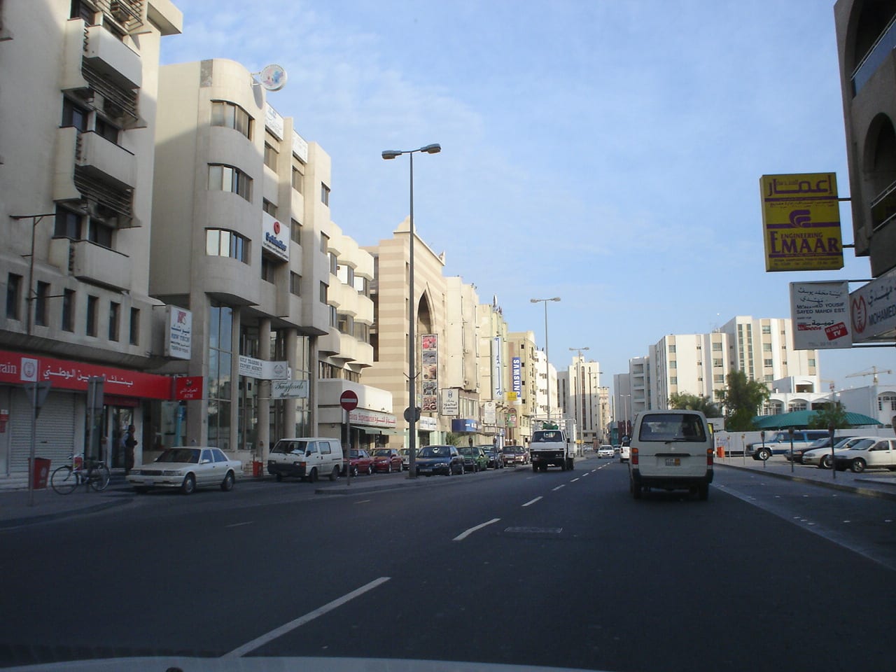 An ordinary Manama street lined with shops and apartment blocks, cars parked along the curb and parked vehicles down both sides