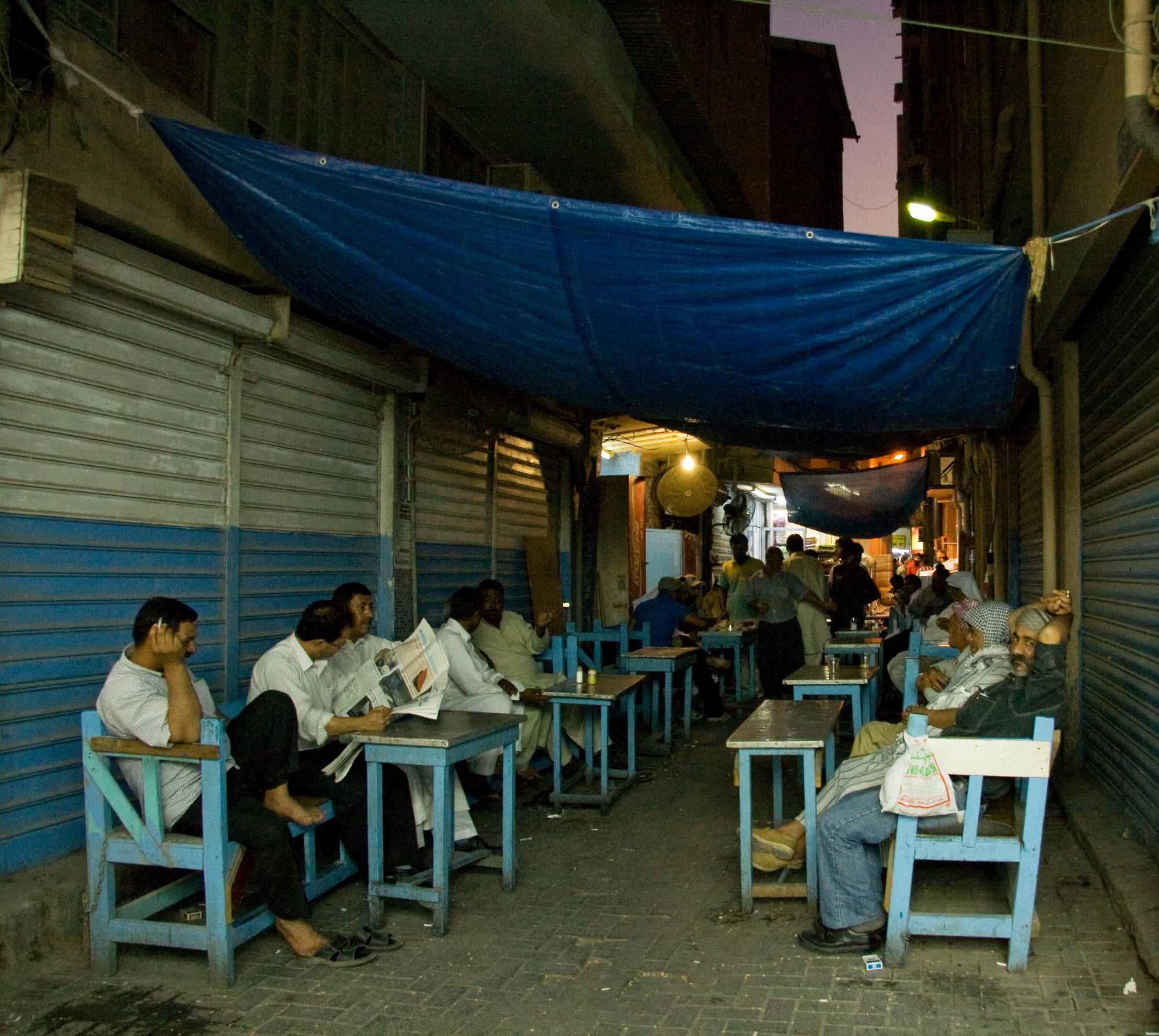 Men sitting at small tables and chairs outside shuttered shopfronts in a narrow souq lane at dusk, reading newspapers and talking