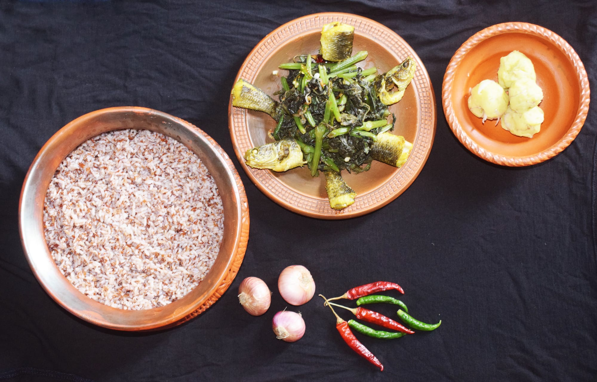 Small bowls of mashed vegetable and fish preparations, greens and fried fish arranged around a bowl of rice