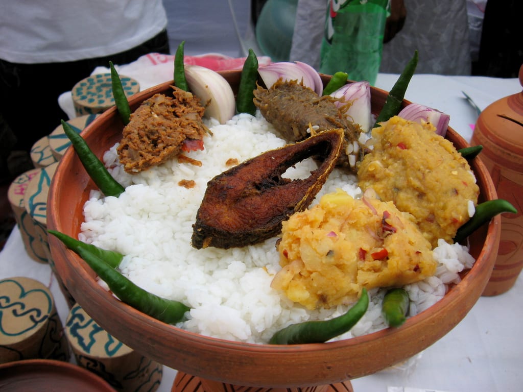A round platter of soaked rice surrounded by small bowls of fried fish, dried fish, dal, pickle, green chillies and onion
