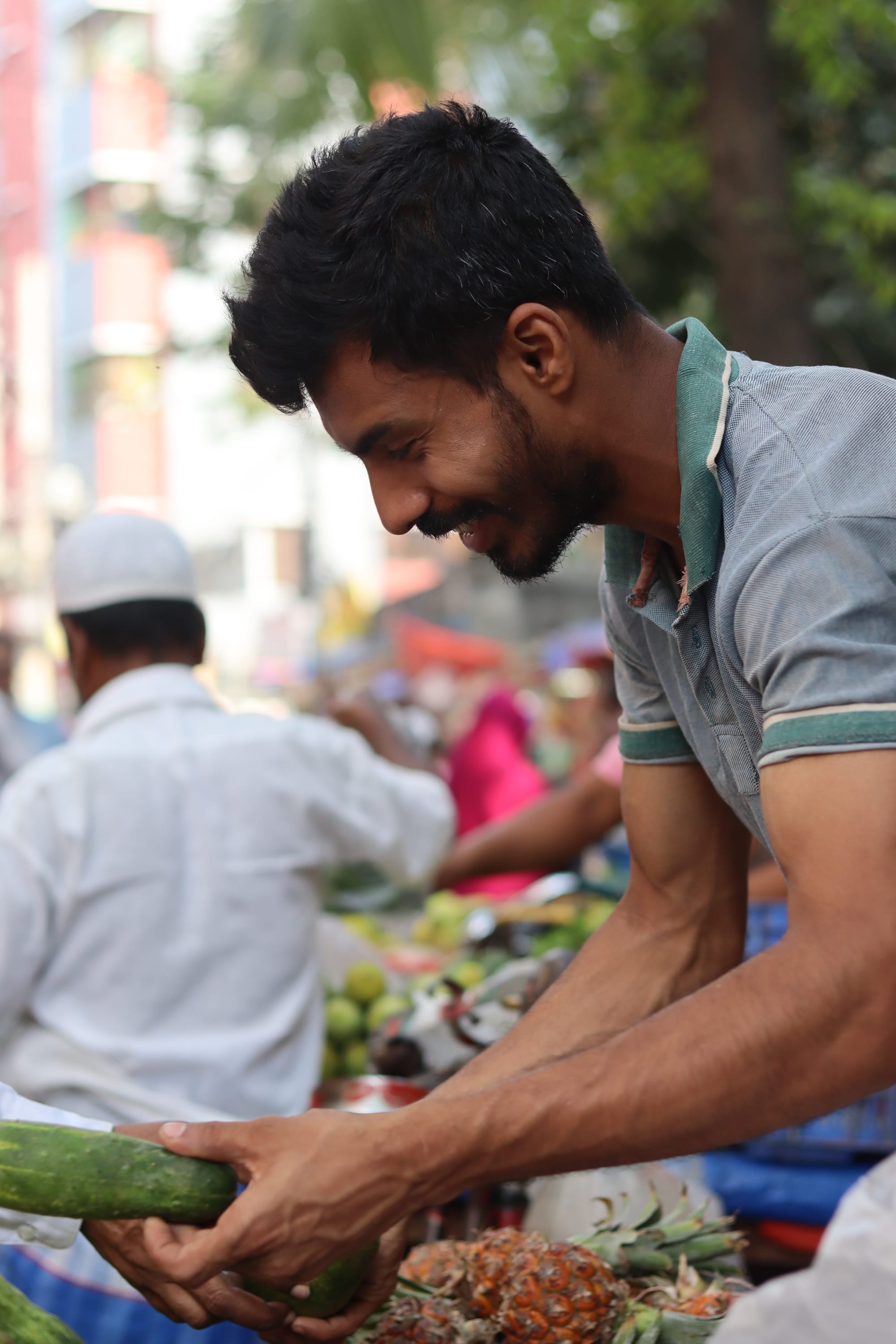 A man at an outdoor market stacking produce, with other vendors and shoppers visible behind him