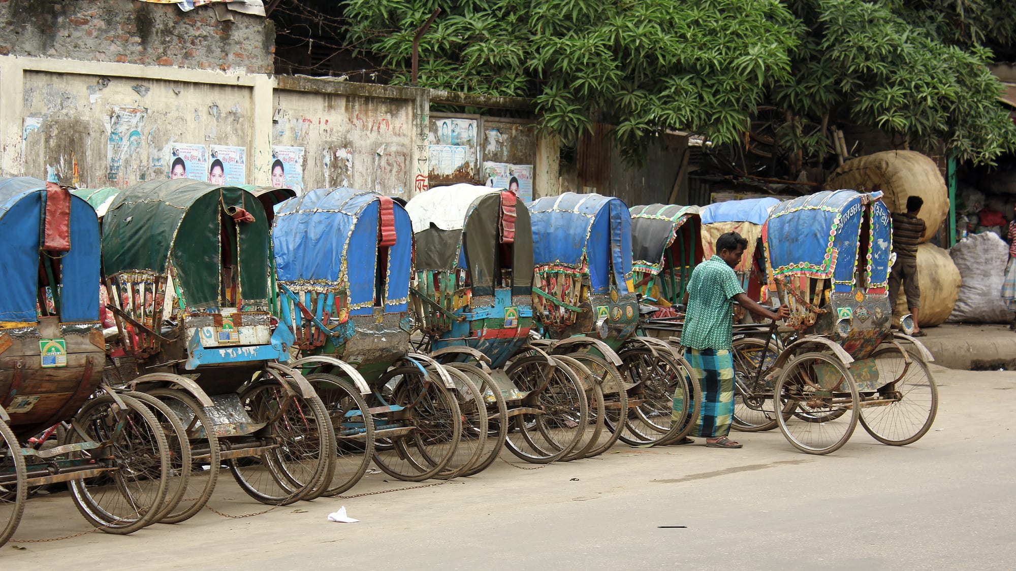 A long row of colourfully decorated cycle rickshaws parked along a street, with one man standing among them