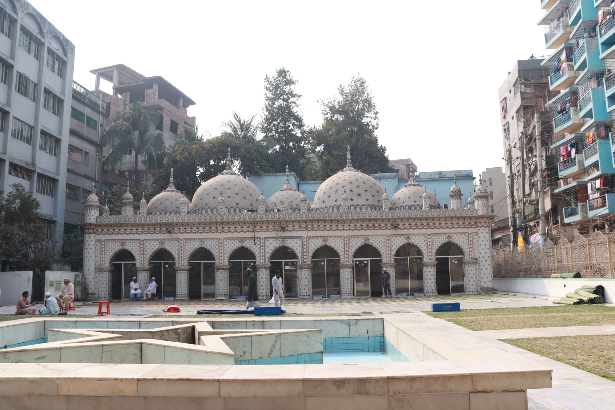 A white mosque with five domes decorated with blue star patterns and a row of arched doorways, seen across a star-shaped pool and paved courtyard, with apartment blocks on either side