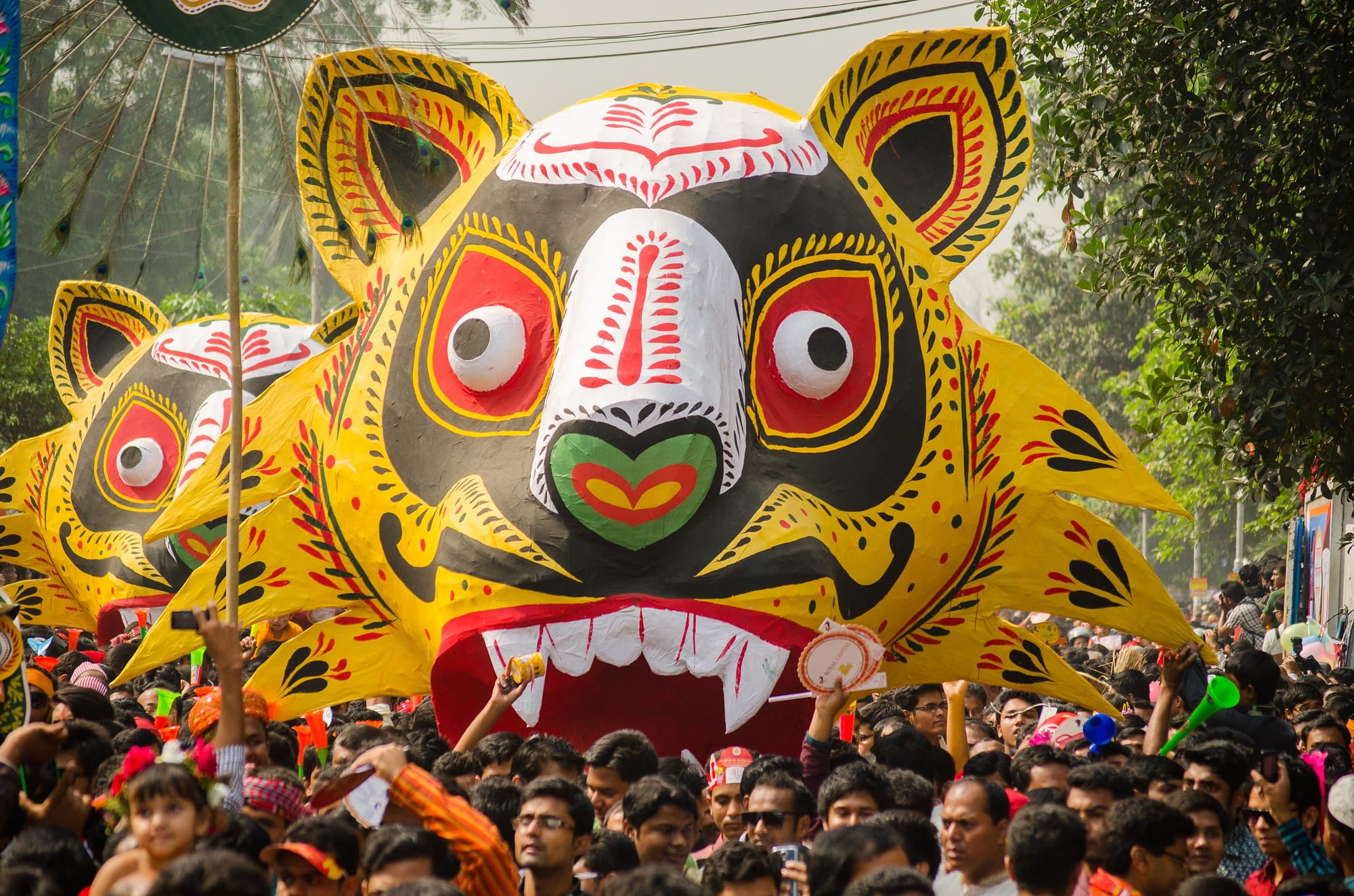 A giant yellow, black and red tiger mask with bulging white eyes and bared teeth carried above a dense crowd, with a second tiger mask behind it