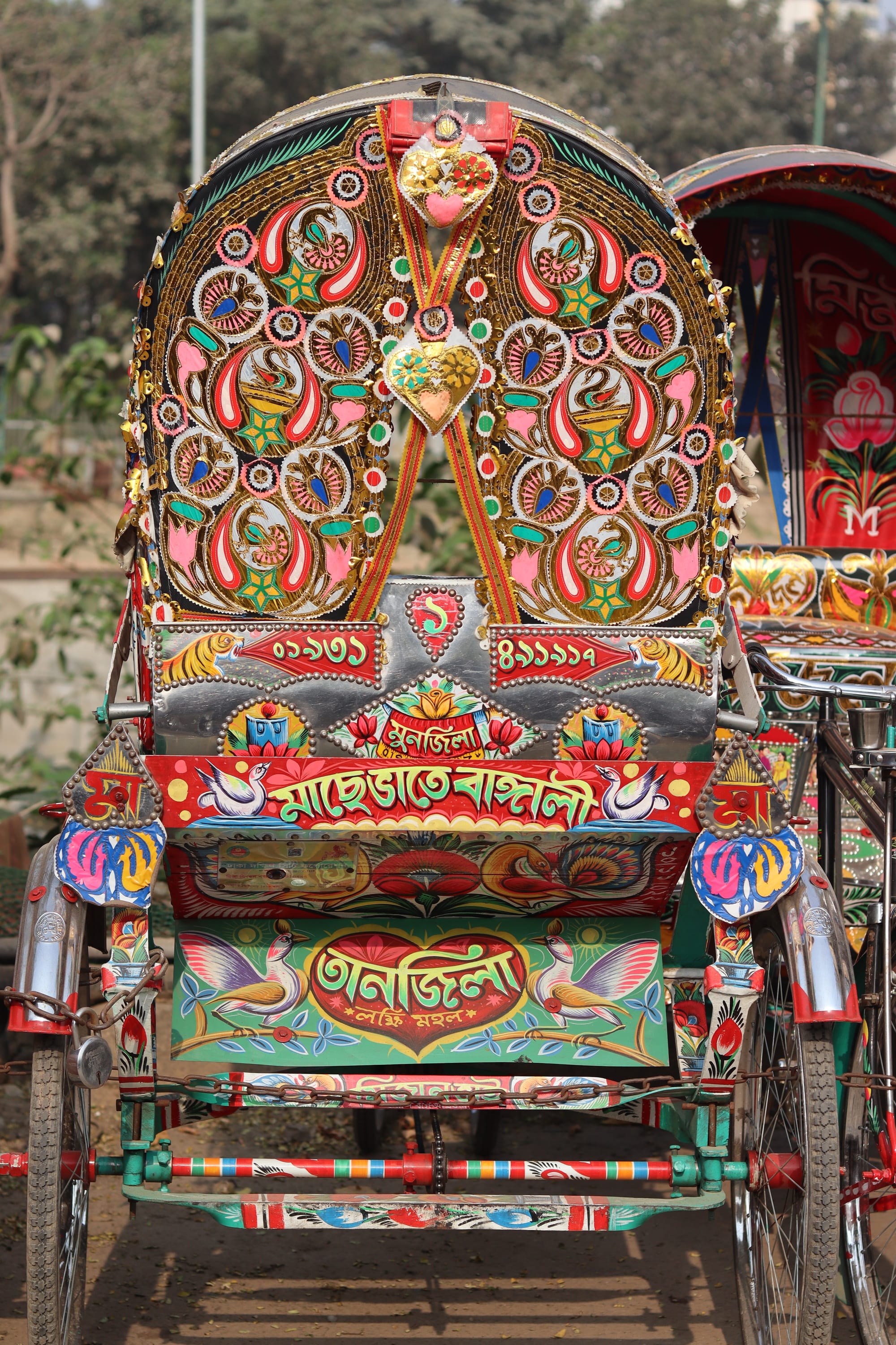 The back of a cycle rickshaw covered in brightly painted decoration, with peacock and paisley motifs on the hood, painted tigers and birds, and Bengali lettering on red and green panels