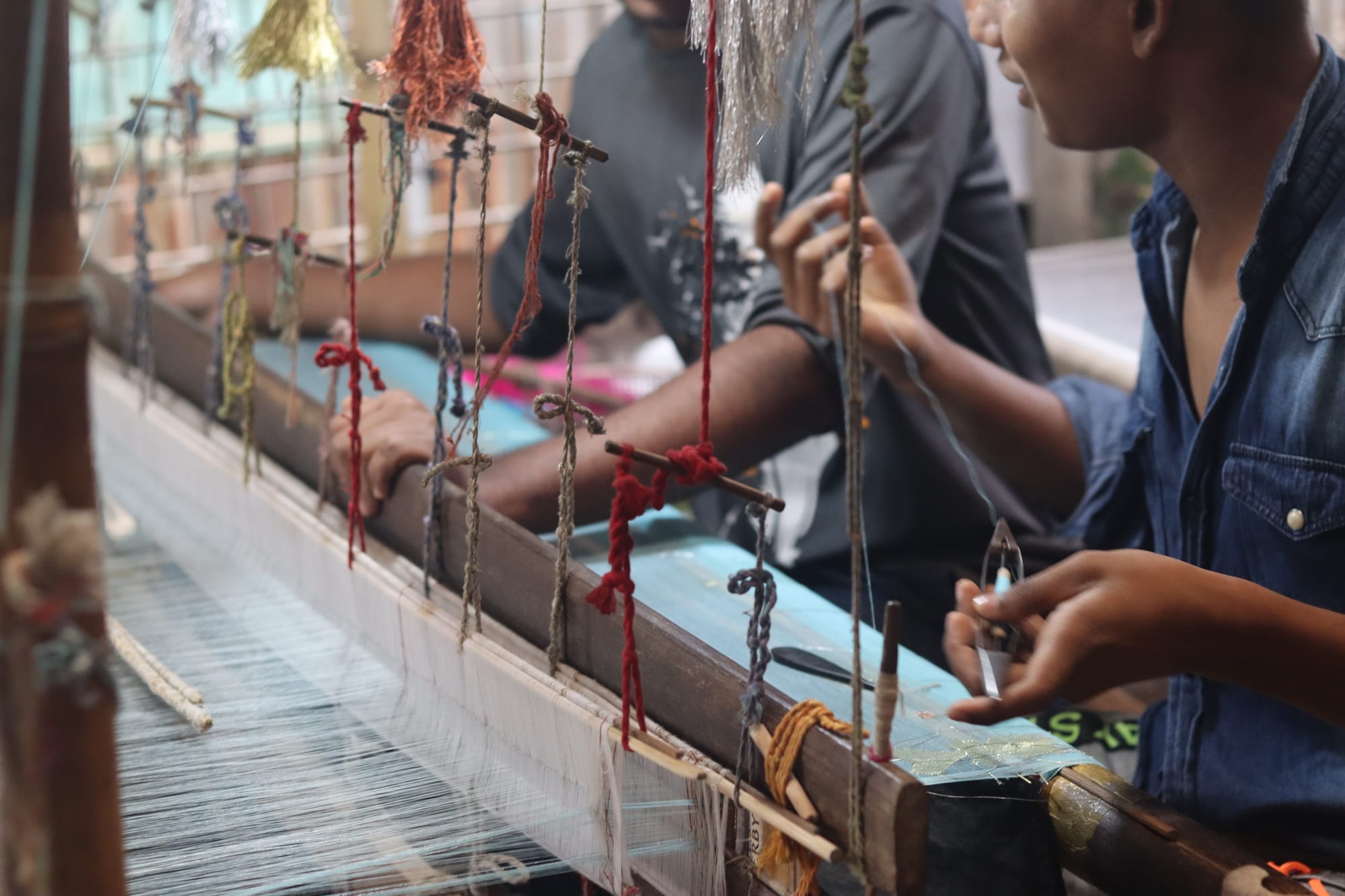 Close view of two weavers' hands working at a wooden handloom, with fine white warp threads, small bobbins of coloured thread and tassels hanging from the loom