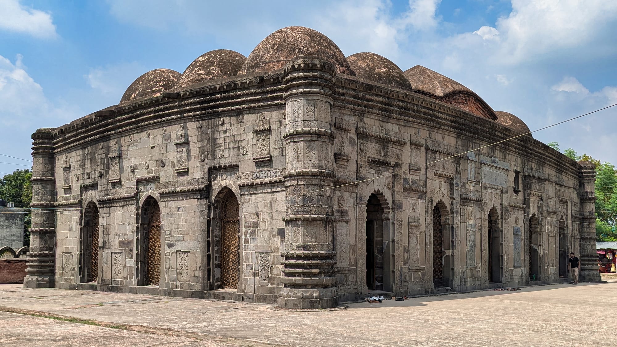 A low rectangular mosque faced in dark carved stone, with arched doorways, round corner towers and a row of weathered domes