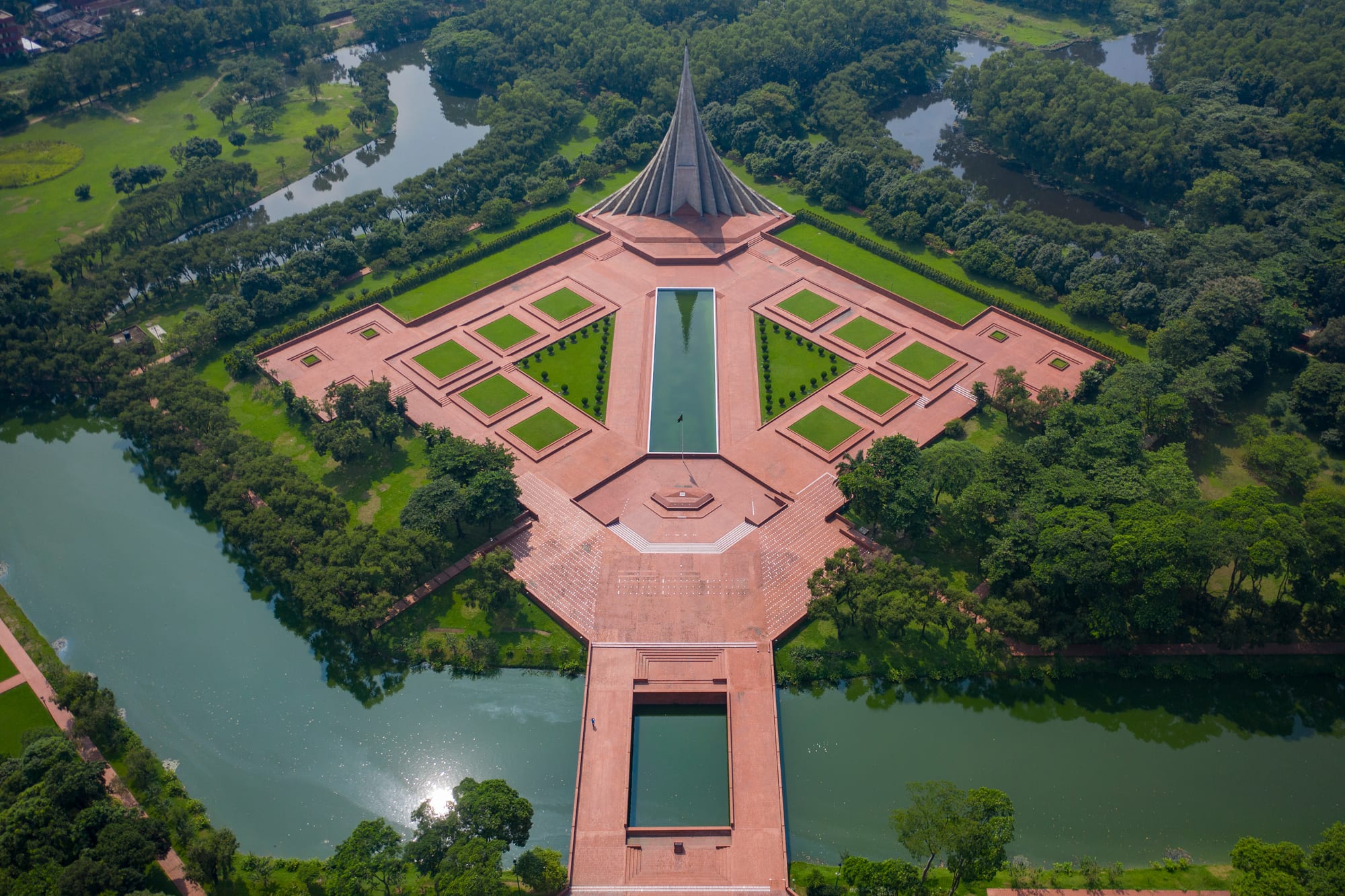 An aerial view of a tall concrete monument of nested pointed triangles at the head of a red-brick plaza with a long reflecting pool, surrounded by trees and water