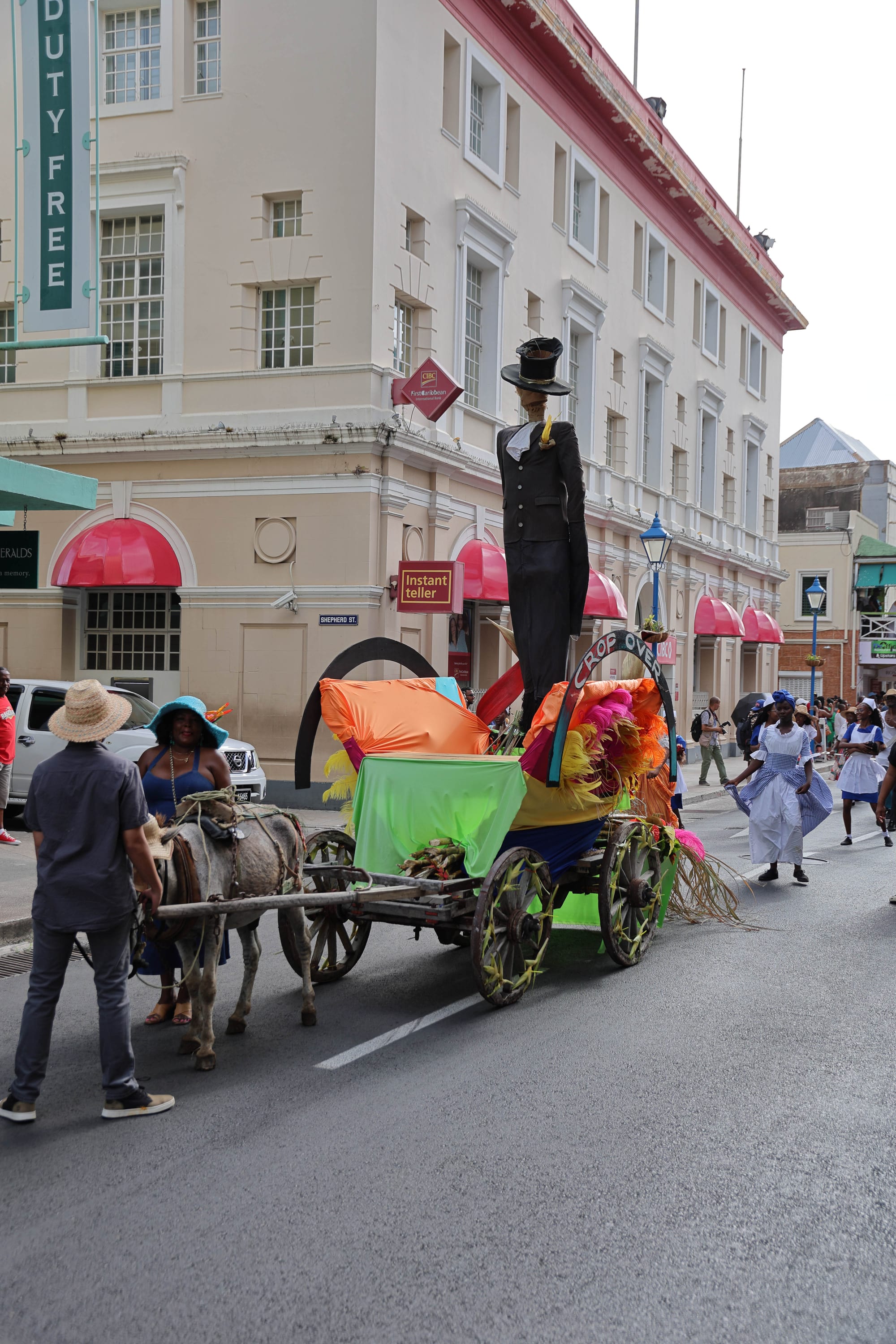 A donkey pulls a wooden cart draped in bright orange, green and blue cloth, feathers and sugar cane, topped by a tall effigy in a black top hat and tailcoat and a sign reading Crop Over, with dancers in blue and white dresses behind