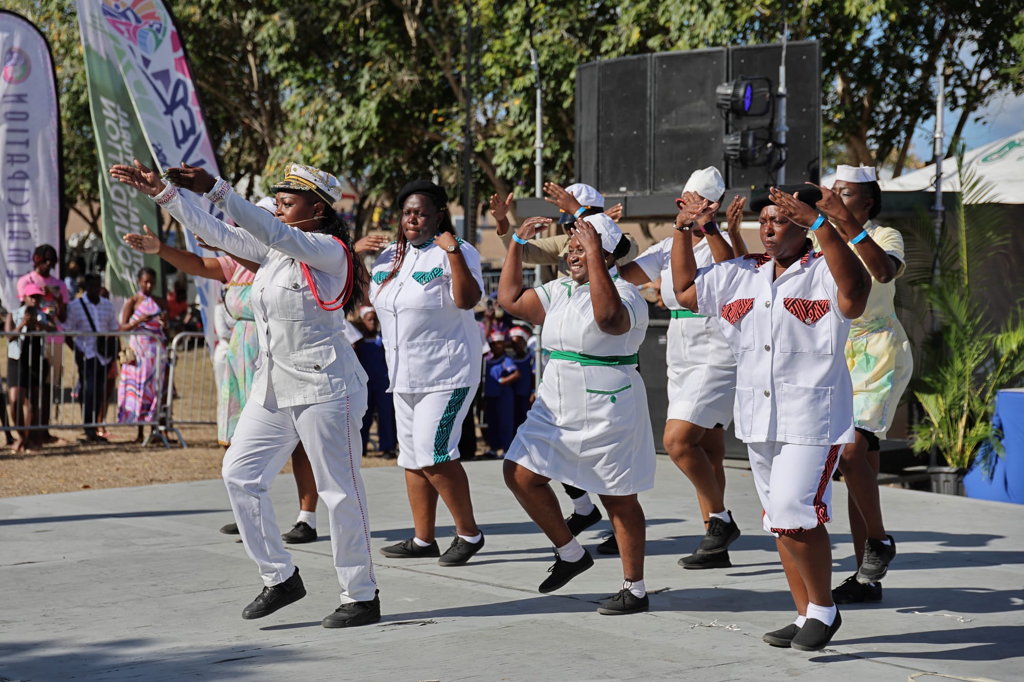 A group of women in white naval-style uniforms with sailor caps and coloured trim dance on an open-air stage, arms outstretched, led by a woman in an officer's cap with red braid