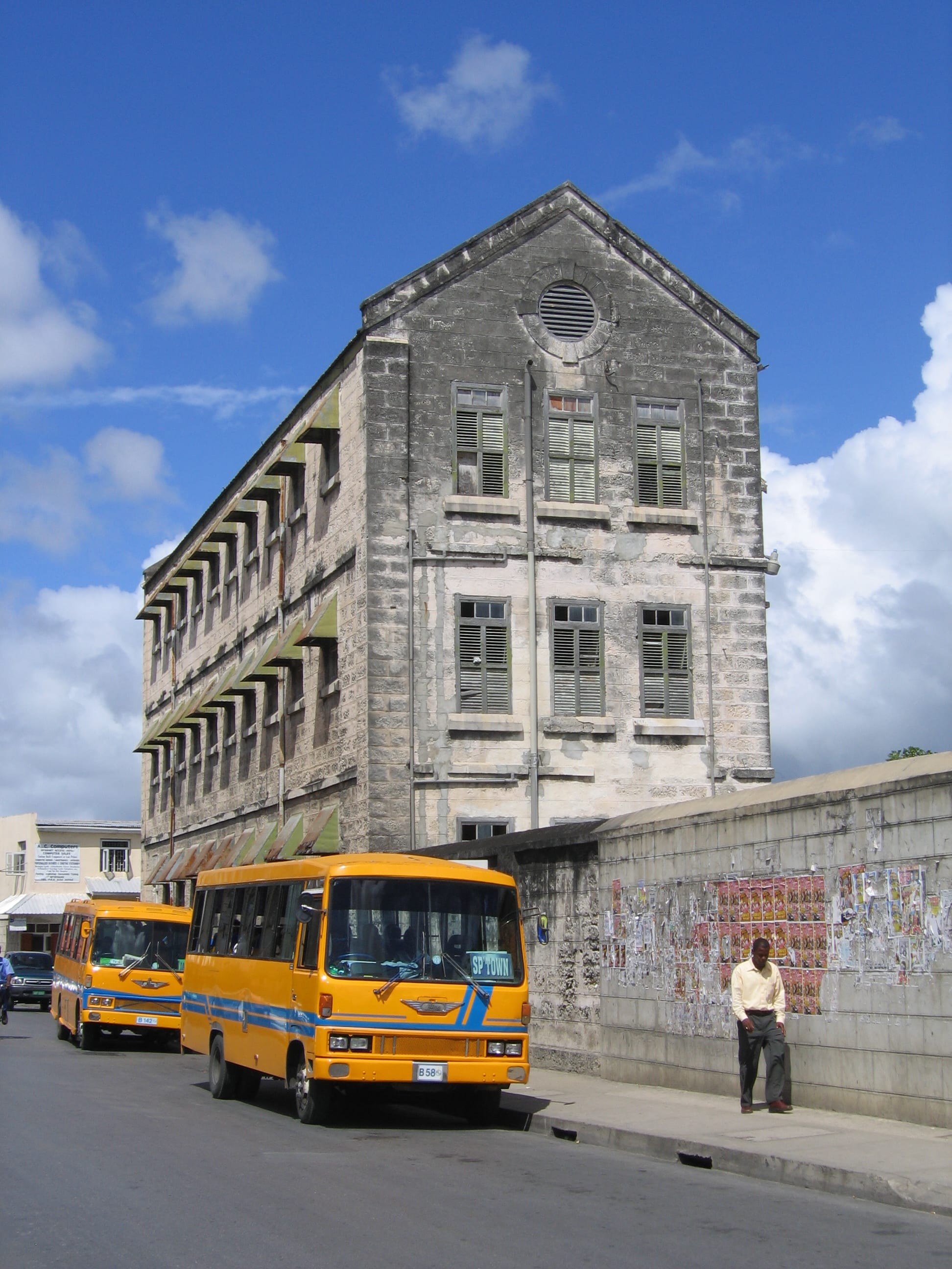 Two yellow privately run minibuses parked on a Bridgetown street below a weathered stone building, with a man waiting on the pavement