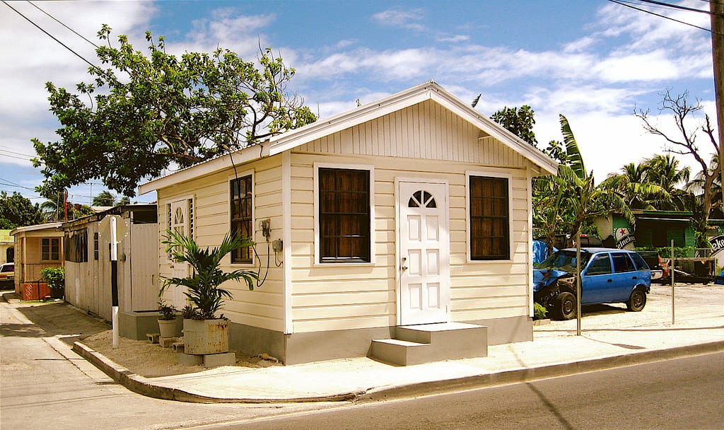 A cream-coloured wooden chattel house on a street corner, with a car parked beside it and palm trees behind