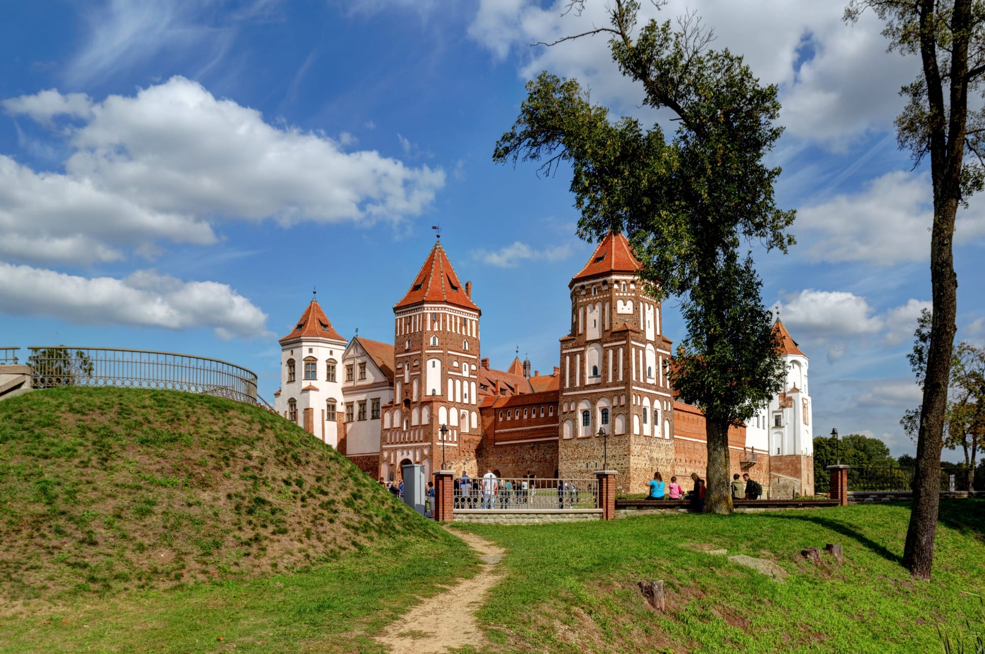 A red-brick castle with tall round corner towers and a Renaissance palace wing, reflected in a pond