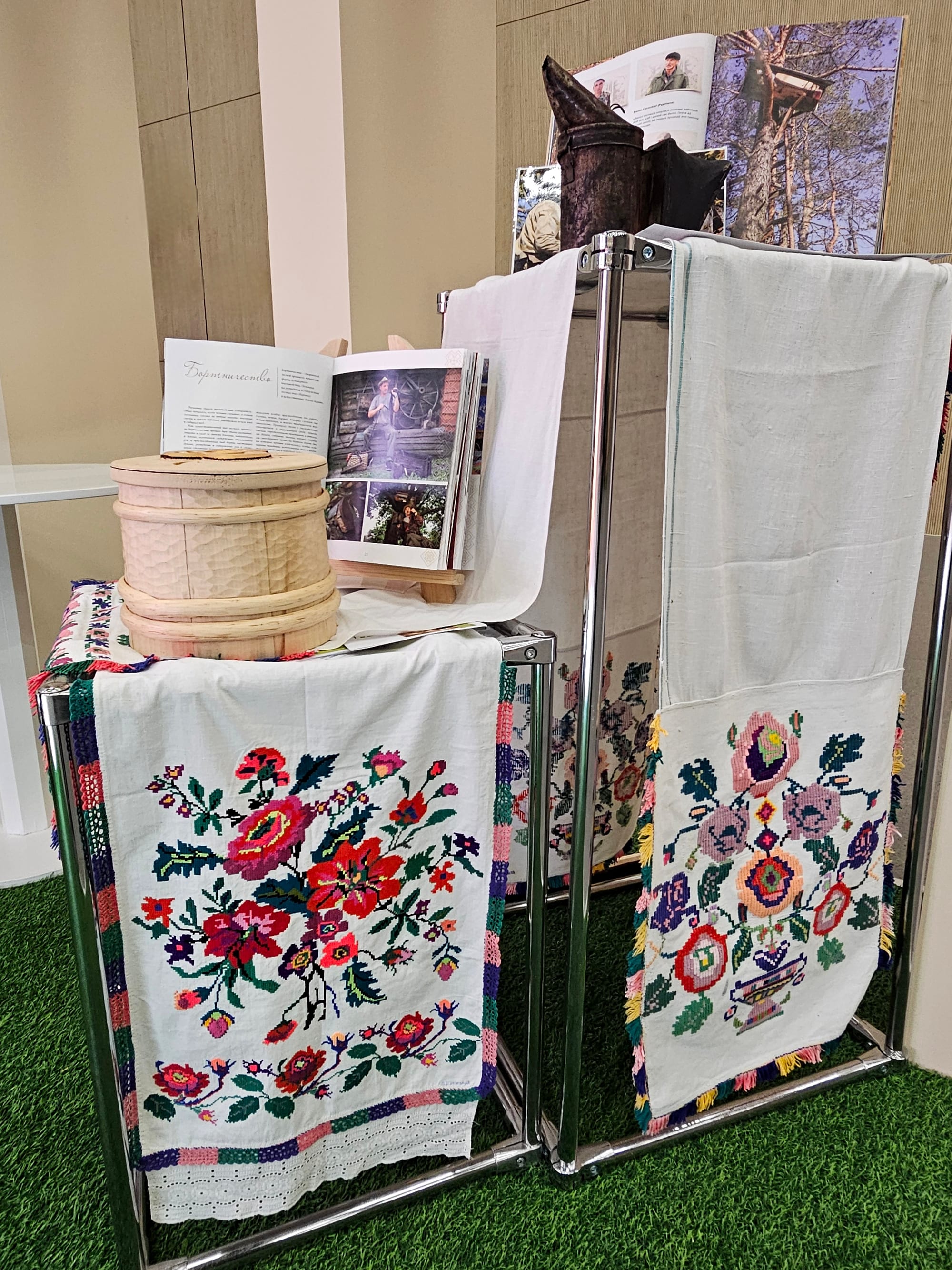 Two long white linen towels embroidered with large red, pink and purple flowers and fringed borders, hanging over metal stands beside a carved wooden container