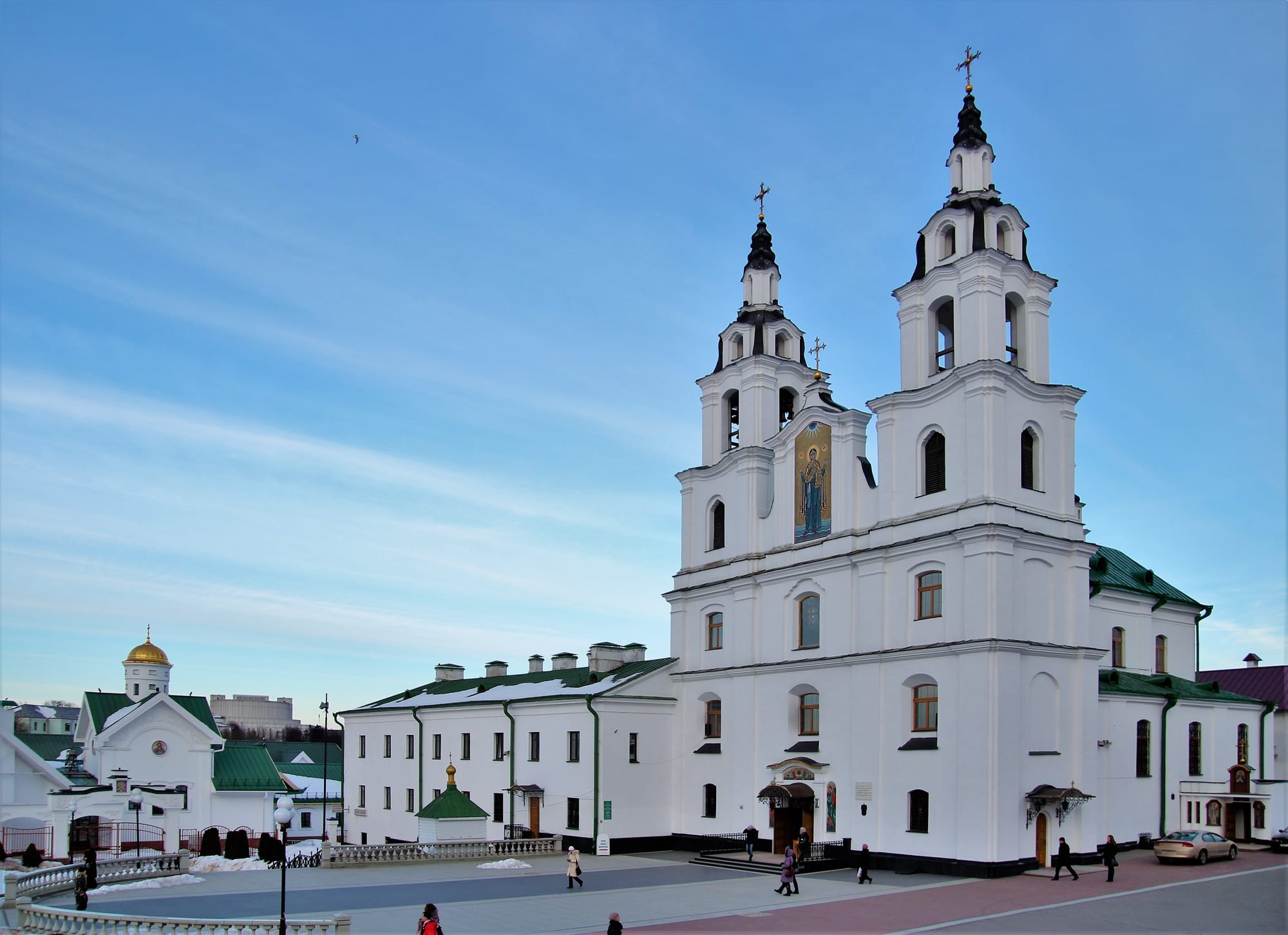 A white Baroque cathedral with twin bell towers topped by crosses and green roofs, an icon set into the facade between the towers, under a clear blue sky